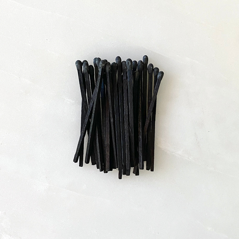 A bunch of handmade black match sticks from House of Good wooden matchbox on a white surface.