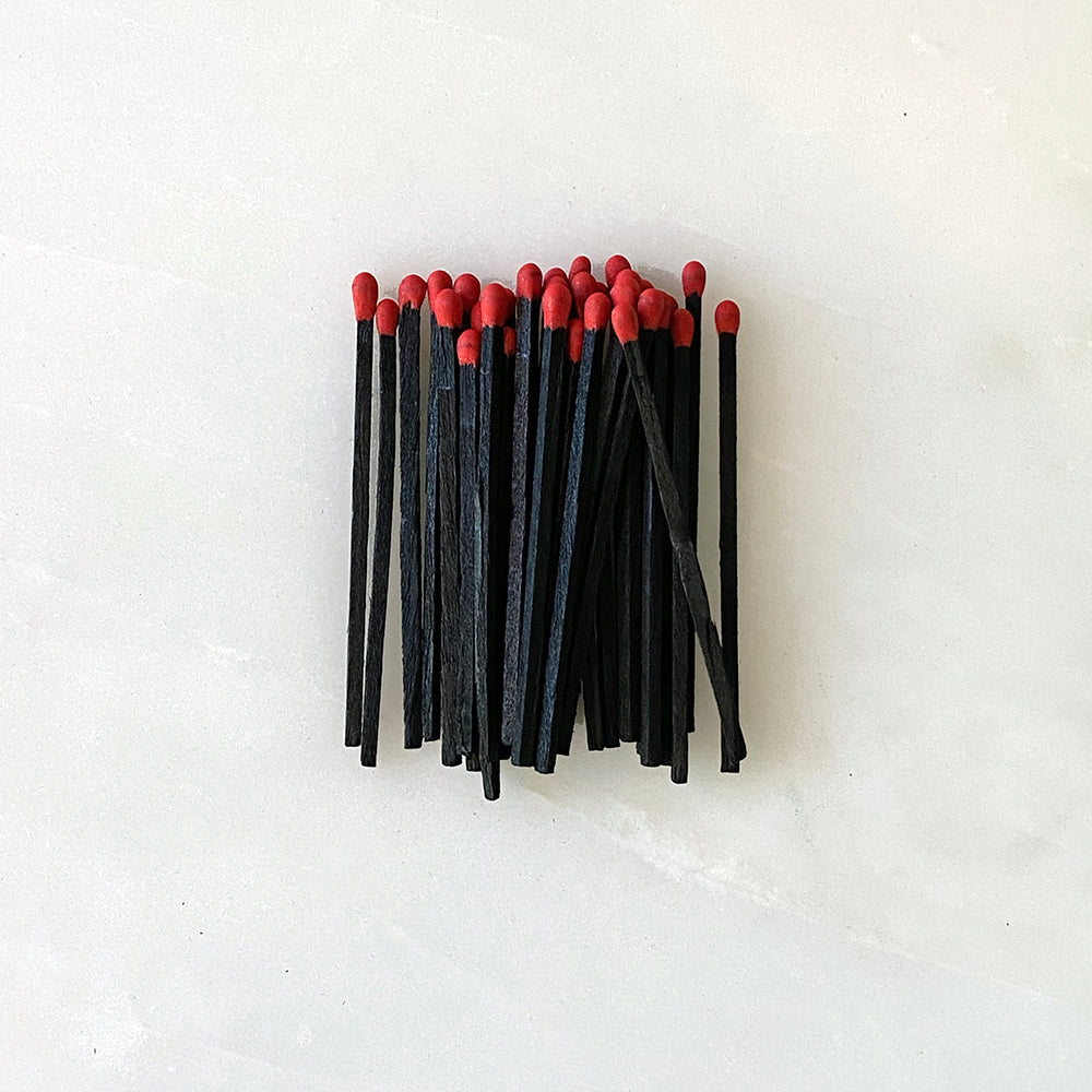 A bunch of handmade black and red matches on a white surface, next to House of Good wooden matchboxes with the House of Good shield.