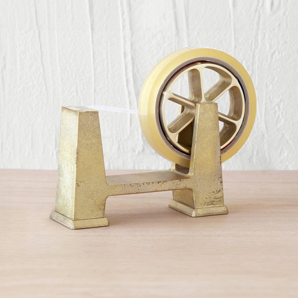 The Futagami Ihada Tape Dispenser, made of solid brass, showcases both beauty and functionality on a wooden table.