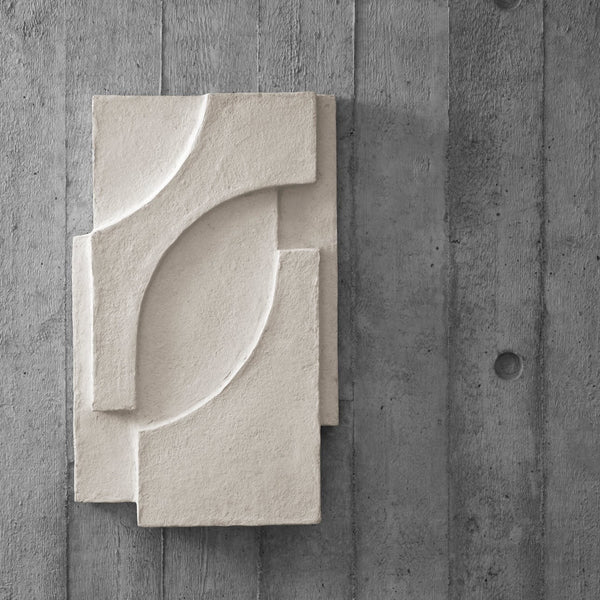 A textured white piece of art, the SERIF RELIEF by KRISTINA DAM STUDIO, on a concrete wall.