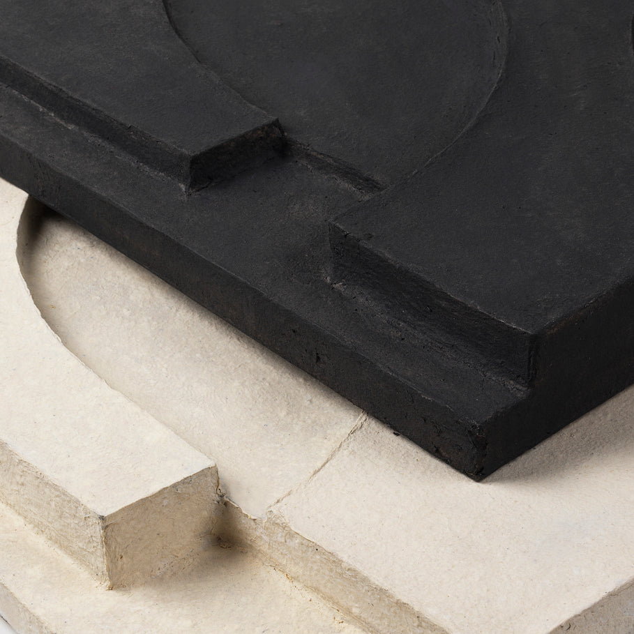 A piece of SERIF RELIEF by KRISTINA DAM STUDIO, a black concrete with textured surfaces, sitting on top of another piece of concrete.