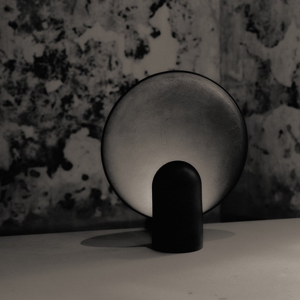 A black and white photo of a SURFACE SCONCE BRASS table lamp by STUDIO HENRY WILSON emitting ambient light.