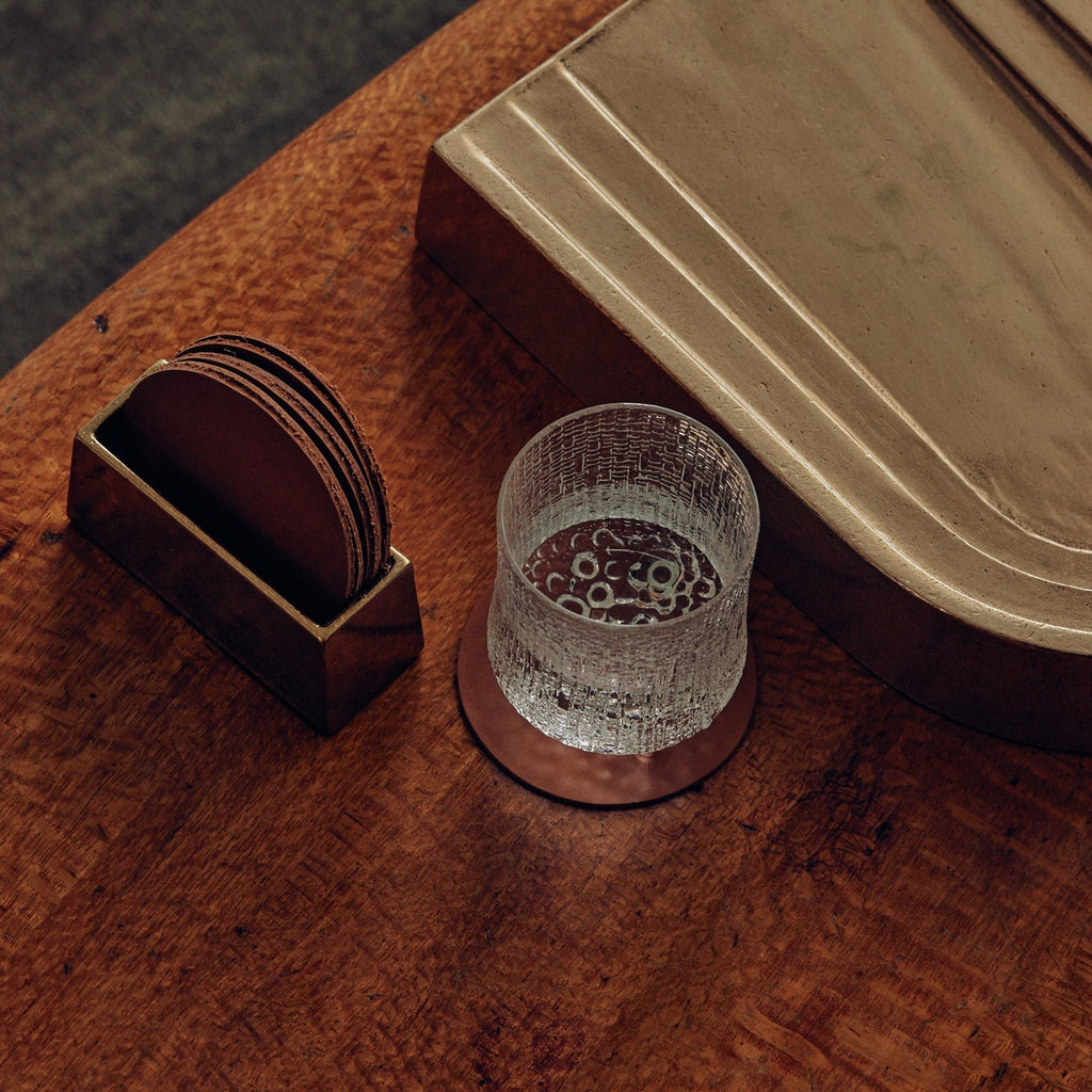 A glass sits on a wooden table beside a Studio Henry Wilson coaster holder with coasters.