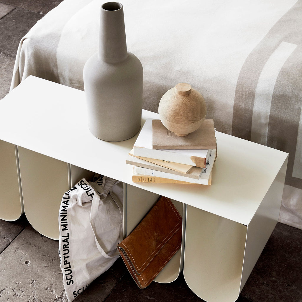 A curved white bedside table by Kristina Dam Studio adorned with a vase and books.