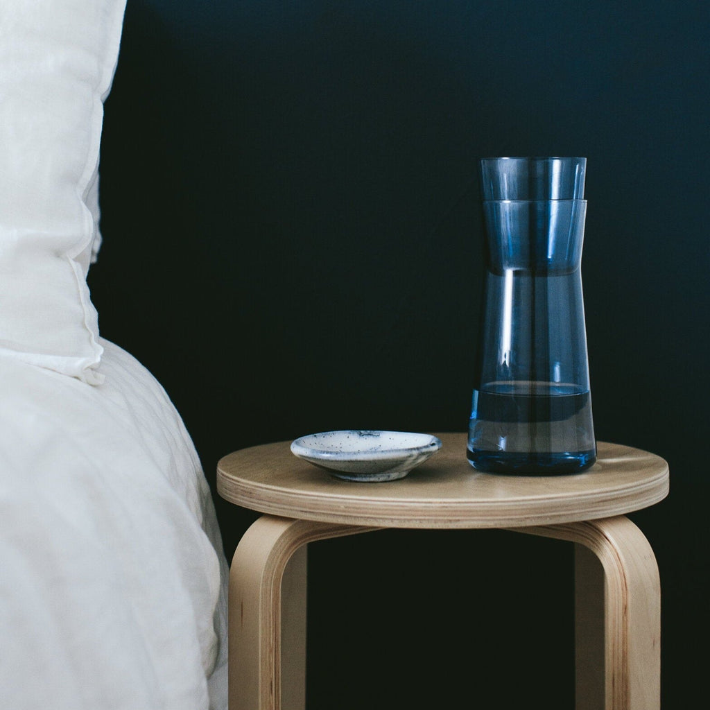 A GLASS CARAFE by GARY BODKER DESIGNS on a wooden table next to a bed, creating a cozy Gestalt Haus ambiance.