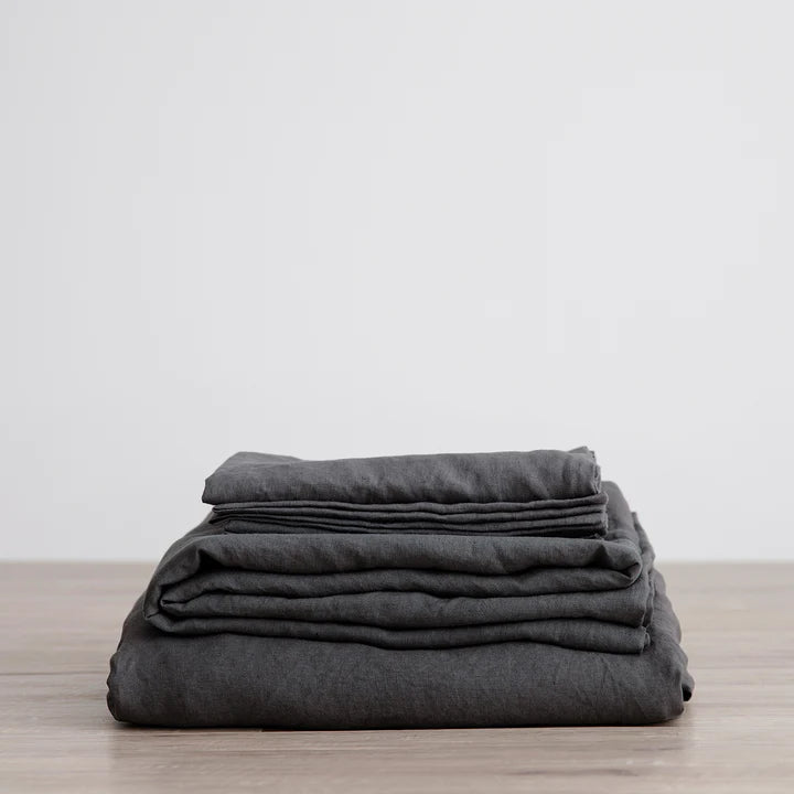 A stack of CULTIVER Linen Sheet Set (Fitted + Flat) with Pillowcases on a wooden table in a Gestalt Haus style.