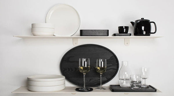 A white Gestalt Haus shelf with black PASSE-PARTOUT coasters by Vincent Van Duysen plates, glasses, and a bottle of wine.