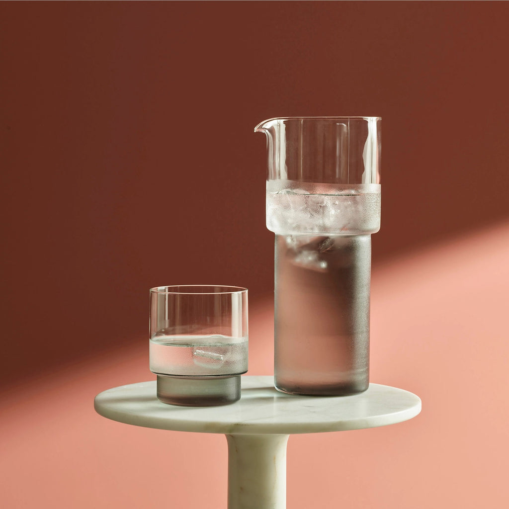A RYE GLASSWARE COLLECTION pitcher and a glass on a table at Gestalt Haus next to a pink wall.